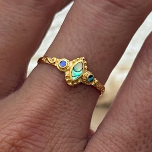 May include: A gold-colored ring with an ornate design. The ring features a central teardrop-shaped abalone shell, flanked by two small blue gemstones. The ring is worn on a finger.