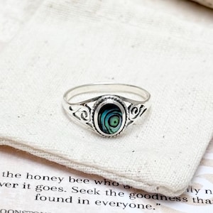 May include: A silver ring featuring an oval abalone shell inlay with iridescent green and blue tones, set within a decorative filigree band. The ring rests on a small off-white fabric pouch placed atop a card with printed text about honey bees.