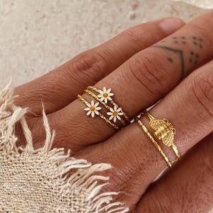 Puede incluir: Anillos dorados con diseños de margaritas y abejas. Un anillo presenta tres margaritas con pétalos blancos y centros amarillos. Otro anillo tiene un diseño de abeja. Los anillos están en una mano.