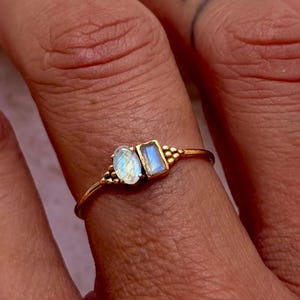 Puede incluir: Un anillo de tono dorado que presenta dos pequeñas piedras preciosas distintas colocadas una al lado de la otra. Una piedra tiene forma ovalada con un aspecto lechoso iridiscente, mientras que la otra es una piedra rectangular de color azul claro.