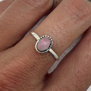 May include: A silver ring with an oval, light pink gemstone. The gemstone is surrounded by a beaded border. The ring is on a finger, with a neutral background.