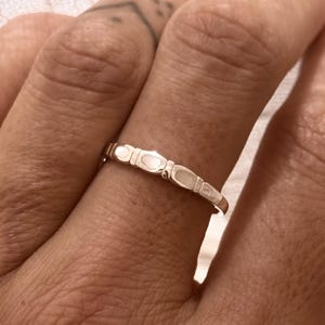May include: A silver ring with a repeating oval pattern. The ring is on a finger, set against a soft, white fabric backdrop. The ring's design consists of small, elongated ovals, giving it a delicate and elegant appearance. The ring is a piece of jewellery.