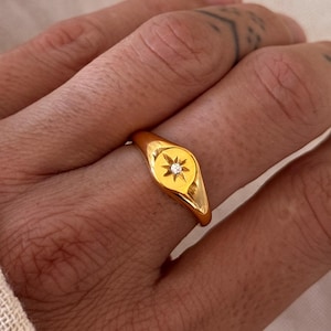 May include: A gold signet ring with a starburst motif and a small, clear gemstone. The ring is worn on a finger, set against a backdrop of soft, neutral-toned fabric. The ring's design is simple and elegant, suitable for everyday wear.