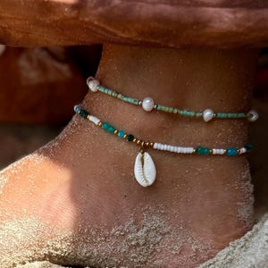 Könnte beinhalten: Zwei Perlen-Fußkettchen mit Strand-Thema. Ein Fußkettchen ist türkis und weiß mit einem kleinen Muschelanhänger. Das andere Fußkettchen ist türkis und weiß mit kleinen Perlen. Die Fußkettchen werden an einem mit Sand bedeckten Knöchel getragen.