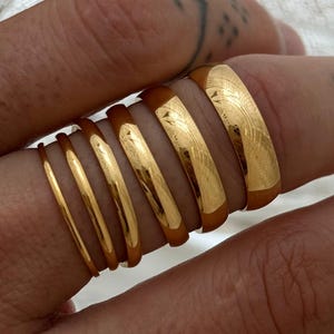 May include: A selection of gold rings in different styles. The rings, worn on a finger, display polished surfaces and detailed designs. Some rings feature etched patterns, while others are plain. The rings are a mix of widths.