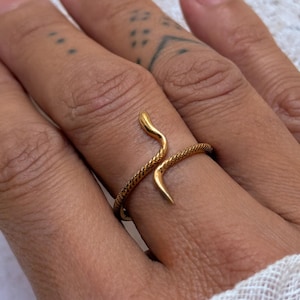 Peut inclure: Une bague dorée en forme de serpent enroulé. L'anneau présente un motif texturé rappelant des écailles sur le corps du serpent, tandis que la tête et la queue sont lisses et effilées. La bague est un modèle ouvert qui entoure le doigt.