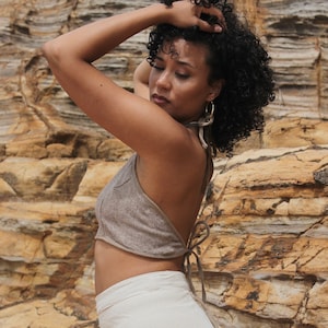 Model standing side on wearing a textured halter neck crop top which ties at the middle of back and back of neck. Light brown mushroom colour Raw cotton woven. Fitted around bust. Model wears halter top with light coloured high waisted wrap skirt.