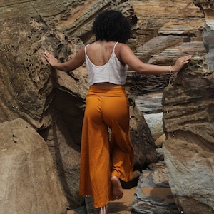 May include: A white sleeveless crop top with thin straps and a textured finish paired with high-waisted, wide-leg trousers in a solid mustard yellow colour. The trousers feature a wide, folded waistband and a loose, flowing fit that reaches the ankles.
