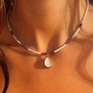 May include: A necklace with a white and marble-effect beaded strand, interspersed with small purple beads and gold-coloured spacers. A teardrop-shaped pendant with a white stone hangs from the centre, suspended by a gold-coloured bail. The necklace is worn around the neck.
