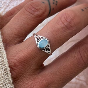 Könnte beinhalten: Ein silberner Ring mit einem ovalen, hellblauen Edelstein. Der Ring hat ein dekoratives filigranes Design um den Edelstein herum. Der Ring befindet sich an einem Finger.