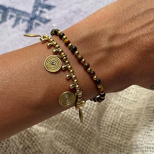 May include: Two bracelets worn together. One bracelet features small brown and black tiger eye beads. The second bracelet consists of metallic gold-toned beads with dangling gold-toned charms, including two circular spiral pendants and small teardrop shapes.