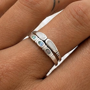 May include: Two silver rings stacked on a finger. The top ring has three small, oval-shaped inlays, one blue, one white, and one grey. The bottom ring is a simple band. The rings are on a hand with a small tattoo.