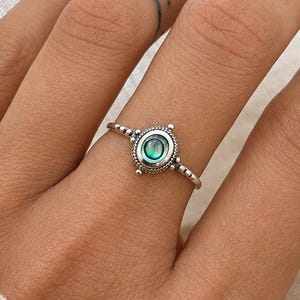 May include: A silver ring featuring an oval, teal gemstone at its centre. The ring has a beaded pattern around the stone and along the band. The ring is worn on a finger, with a tattoo visible on the adjacent finger.