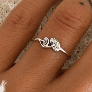 May include: A silver ring with a mushroom design. The ring features two mushroom-shaped elements joined by a slender band. The ring is worn on a finger, set against a textured, light-coloured background.