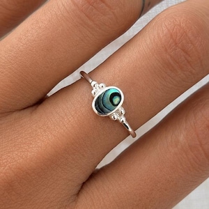 Könnte beinhalten: Ein silberner Ring mit einem ovalen, schillernden Abalone-Muschel-Mittelstein. Der Ring hat ein zartes Band und dekorative Akzente auf beiden Seiten des Steins. Die Abalone-Muschel zeigt Grün-, Blau- und Schwarztöne.