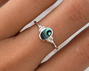 May include: A silver ring with an oval, iridescent abalone shell center stone. The ring has a delicate band and decorative accents on either side of the stone. The abalone shell displays shades of green, blue, and black.