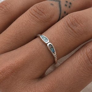 May include: A silver ring featuring two small, teardrop-shaped inlays of blue-green stone. The band has a polished metallic finish with subtle etched line details on the sides, creating a simple and refined appearance for this piece of jewellery.