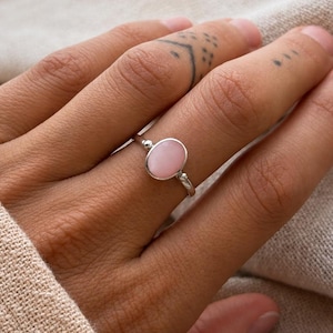 Puede incluir: Un anillo de plata con una piedra lisa de color rosa claro en forma ovalada, montada en una banda de metal sencilla. El engaste incluye dos pequeñas esferas de plata a cada lado de la gema central, con un acabado suave y mate.