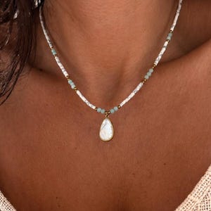 May include: A beaded necklace featuring white marbled cylindrical beads, small light green round beads, and gold-toned spacers. The necklace includes a central teardrop-shaped pendant with a white iridescent inlay set in a gold-toned metal frame.