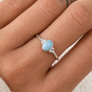 Puede incluir: Un anillo de plata con una piedra preciosa ovalada de color azul claro. El anillo tiene una banda delicada y pequeños detalles decorativos a cada lado de la piedra preciosa. El anillo se muestra en una mano.