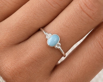 May include: A silver ring featuring a light blue oval gemstone. The ring has a delicate band and small decorative accents on either side of the gemstone. The ring is shown on a hand.