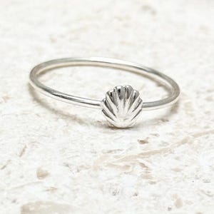 May include: A silver ring featuring a small seashell design. The ring is set against a neutral background, highlighting the delicate details of the seashell. The ring's band is thin and simple, emphasizing the seashell centerpiece.