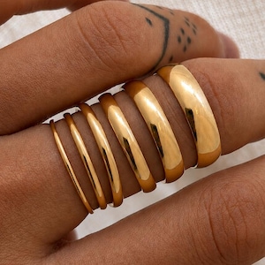 May include: A selection of gold rings of different thicknesses are shown on a finger. The rings vary from slender bands to a broader, rounded style. The rings have a polished, shiny finish, reflecting light. The overall aesthetic is minimalist and elegant.