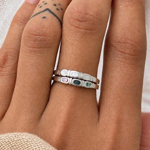 Puede incluir: Dos anillos de plata con diseño ovalado, cada uno con una pequeña piedra de colores. Los anillos están apilados en un dedo, mostrando los delicados detalles y la artesanía de la joyería. Un accesorio sencillo pero elegante.