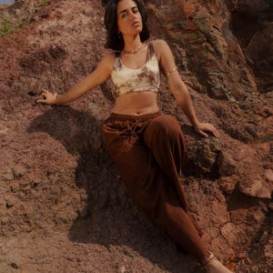 K&ouml;nnte beinhalten: Ein Crop-Top mit Batikmuster in Braun- und Cremet&ouml;nen, kombiniert mit einer braunen Hose mit Kordelzug. Das Model tr&auml;gt Goldschmuck. Das Outfit ist l&auml;ssig und bequem, geeignet f&uuml;r warmes Wetter.