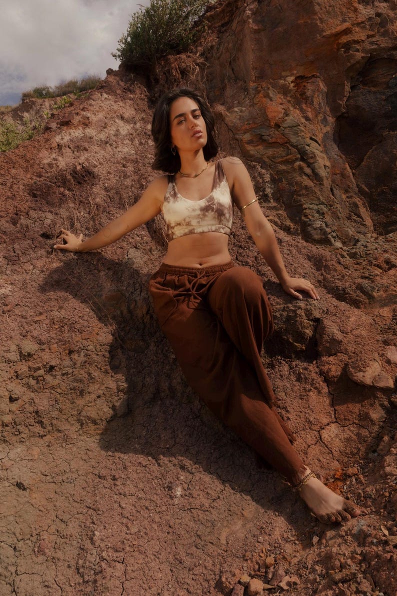 K&ouml;nnte beinhalten: Ein Crop-Top mit Batikmuster in Braun- und Cremet&ouml;nen, kombiniert mit einer braunen Hose mit Kordelzug. Das Model tr&auml;gt Goldschmuck. Das Outfit ist l&auml;ssig und bequem, geeignet f&uuml;r warmes Wetter.
