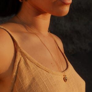 May include: A gold pendant necklace with a small, geometric charm is worn over a tan tank top. The person is also wearing gold hoop earrings. The lighting is warm, creating a soft glow.