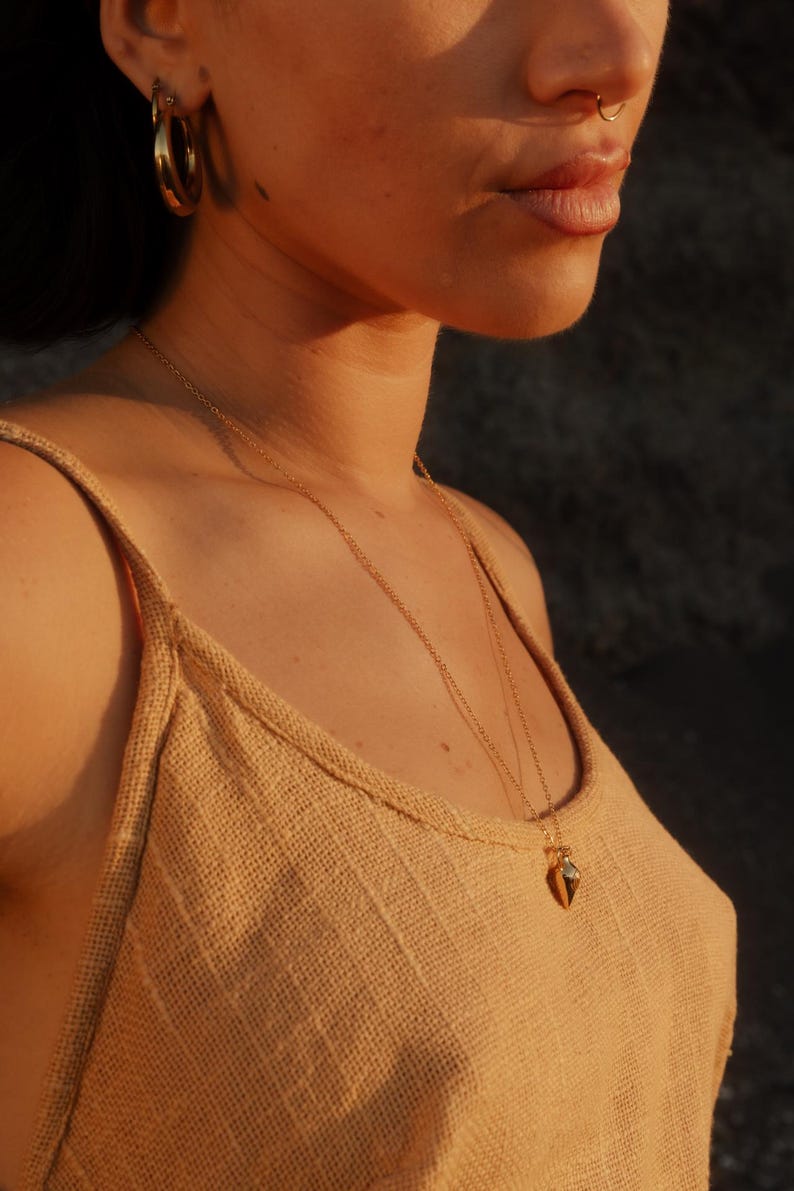 May include: A gold pendant necklace with a small, geometric charm is worn over a tan tank top. The person is also wearing gold hoop earrings. The lighting is warm, creating a soft glow.