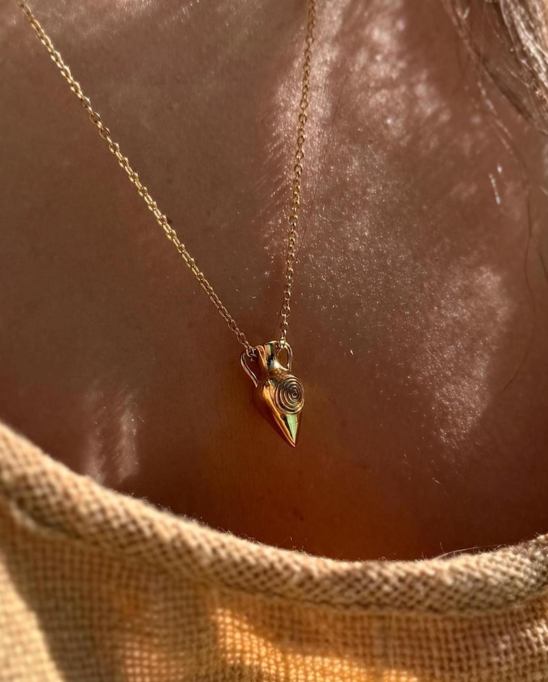 May include: A gold pendant necklace with a small, amphora-shaped charm. The charm has a spiral design and two small handles. The delicate gold chain creates a minimalist look. The necklace is a classic piece of jewellery.