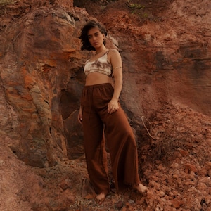 K&ouml;nnte beinhalten: Eine Person tr&auml;gt ein Crop-Top mit Batikmuster und eine braune Hose mit weitem Bein vor einer r&ouml;tlich-braunen Felsformation. Das Outfit hat eine neutrale Farbpalette, wobei das Oberteil Beige- und Braunt&ouml;ne aufweist.