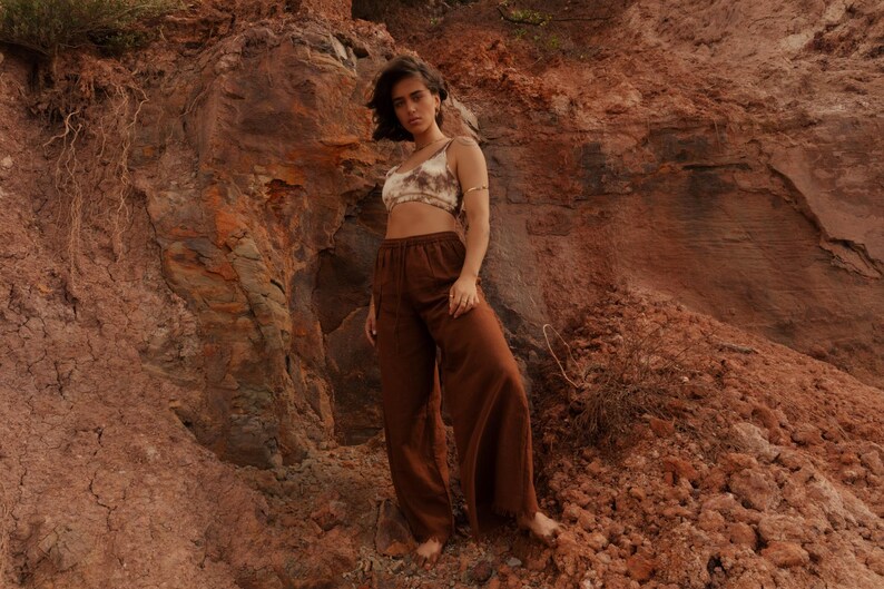 K&ouml;nnte beinhalten: Eine Person tr&auml;gt ein Crop-Top mit Batikmuster und eine braune Hose mit weitem Bein vor einer r&ouml;tlich-braunen Felsformation. Das Outfit hat eine neutrale Farbpalette, wobei das Oberteil Beige- und Braunt&ouml;ne aufweist.
