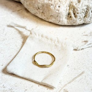 May include: A gold-colored ring with a textured surface rests on a small white drawstring bag. The ring has an organic, irregular shape. The background includes a large, light-colored stone and a textured surface. The text "GIFT READY PACKAGING SHIPS NEXT BUSINESS DAY" is visible.