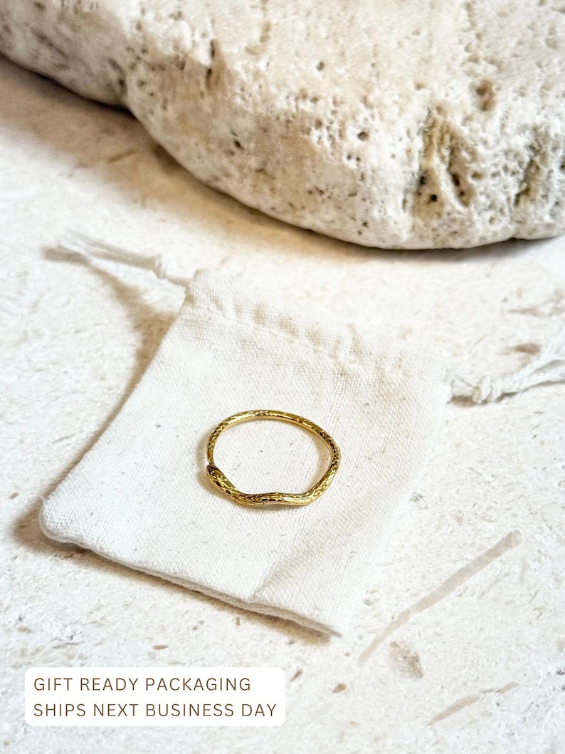 May include: A gold-colored ring with a textured surface rests on a small white drawstring bag. The ring has an organic, irregular shape. The background includes a large, light-colored stone and a textured surface. The text "GIFT READY PACKAGING SHIPS NEXT BUSINESS DAY" is visible.