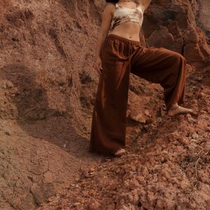 K&ouml;nnte beinhalten: Ein braunes Crop-Top und eine Hose mit weitem Bein. Das Oberteil hat ein Batikmuster in Braun- und Cremet&ouml;nen. Die Hose hat einen Kordelzug in der Taille. Das Model steht auf einer felsigen, rotbraunen Oberfl&auml;che.