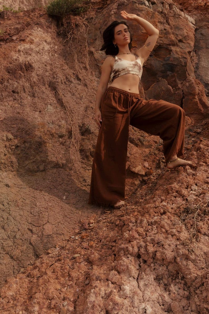 K&ouml;nnte beinhalten: Ein braunes Crop-Top und eine Hose mit weitem Bein. Das Oberteil hat ein Batikmuster in Braun- und Cremet&ouml;nen. Die Hose hat einen Kordelzug in der Taille. Das Model steht auf einer felsigen, rotbraunen Oberfl&auml;che.