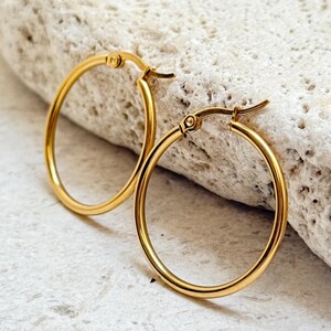 May include: Two gold hoop earrings. The earrings are round and have a shiny finish. The earrings are displayed on a light-coloured, textured surface. The earrings have a simple, classic design.