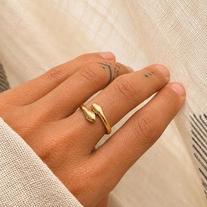 May include: A gold-colored ring in the shape of a snake, wrapped around a finger. The ring has a textured surface and is set against a neutral background. The snake's head and tail meet on the finger.