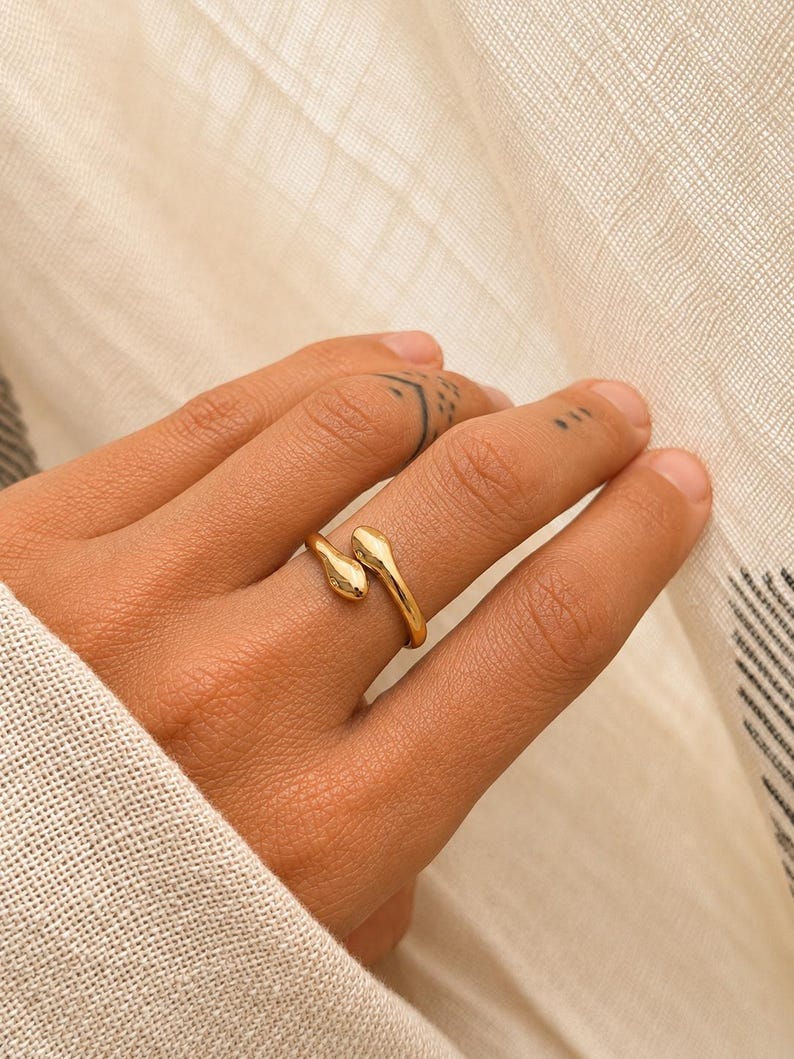 May include: A gold-colored ring in the shape of a snake, wrapped around a finger. The ring has a textured surface and is set against a neutral background. The snake's head and tail meet on the finger.