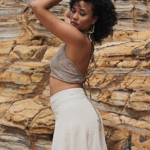May include: A woman in a beige halter top and a cream skirt stands before a rock formation. The halter top ties at the neck and back. The skirt is long and flowing. The woman has curly hair and wears hoop earrings. The outfit is neutral-toned.