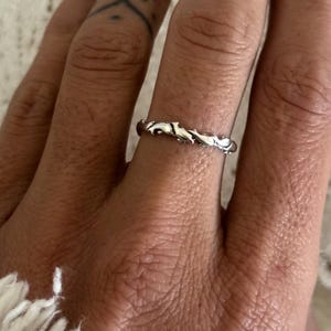 May include: A silver ring with a delicate, textured design. The ring features a repeating pattern of stylized, organic shapes. The ring is worn on a finger, showcasing its intricate details and craftsmanship. The ring is a piece of jewellery.