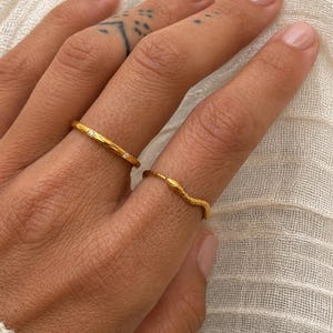 May include: Two gold rings are displayed on a hand. One ring is a thin band with a textured surface and a small diamond. The other ring has a wavy, textured design. The rings are set against a soft, white background.