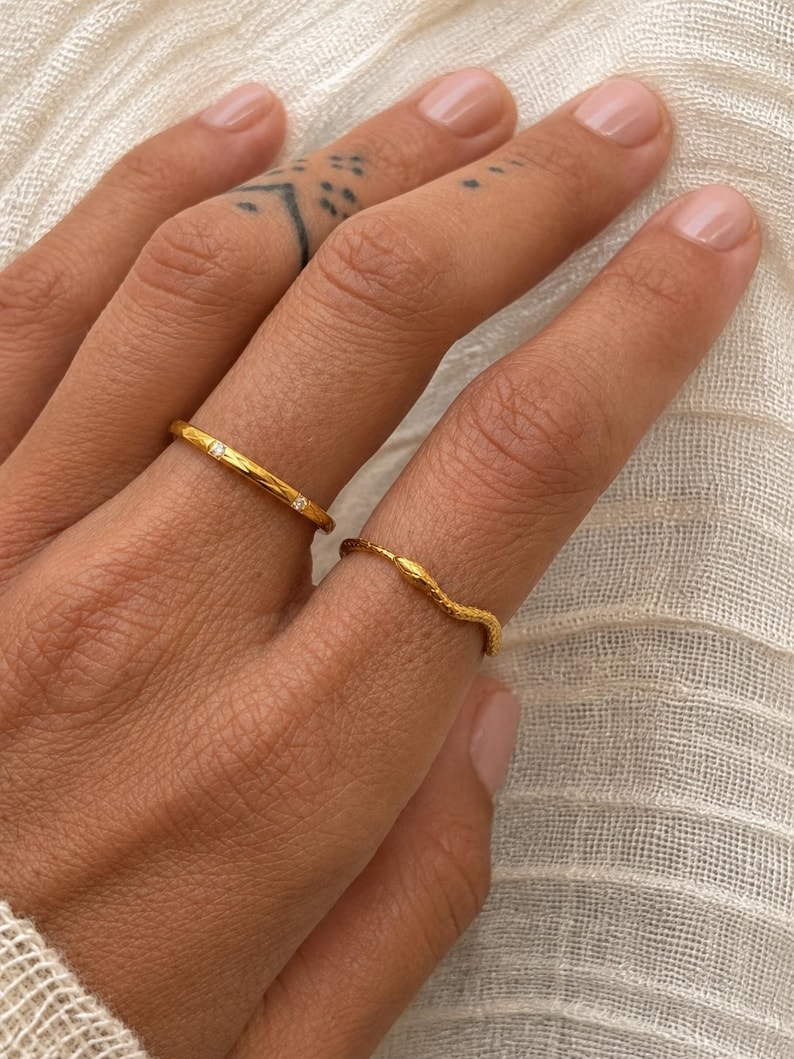 May include: Two gold rings are displayed on a hand. One ring is a thin band with a textured surface and a small diamond. The other ring has a wavy, textured design. The rings are set against a soft, white background.