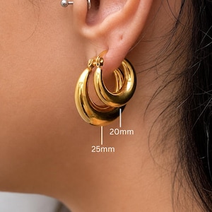 May include: Gold-colored hoop earrings, 20mm and 25mm in size, are shown in this image. The earrings have a smooth, polished finish and are displayed on an ear. The earrings are a classic jewelry item.