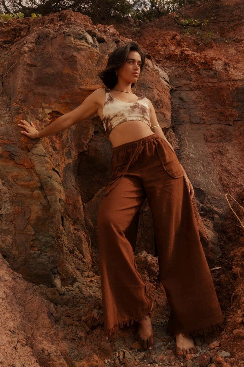 K&ouml;nnte beinhalten: Eine Frau tr&auml;gt ein bauchfreies Oberteil mit Batikmuster und eine braune Hose mit weitem Bein. Die Hose hat einen Kordelzug in der Taille und Fransen am Saum. Das Outfit befindet sich vor r&ouml;tlich-braunen Felsformationen.