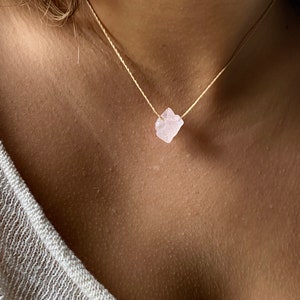 May include: A delicate necklace featuring a raw, light pink rose quartz crystal pendant. The crystal is suspended from a thin, beige cord. The necklace is shown against a skin tone background, highlighting the natural beauty of the gemstone.
