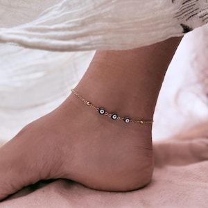 May include: A gold-colored anklet featuring three blue and white evil eye charms. The delicate chain is adorned with small, round beads. The anklet is worn on a foot, with a white fabric in the background.